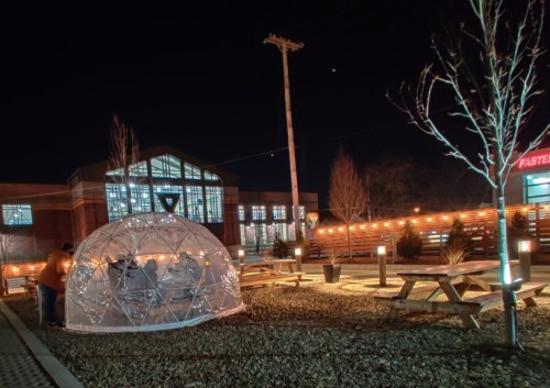 A clear igloo structure in a lit outdoor area at night, surrounded by picnic tables and trees.