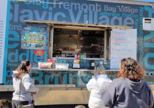 A food truck with a menu displayed, customers waiting in line to order. Bright blue and black design on the truck.