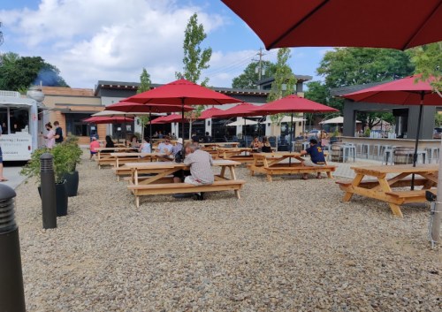 Outdoor dining area with picnic tables and red umbrellas, filled with people enjoying their meals in a gravel setting.