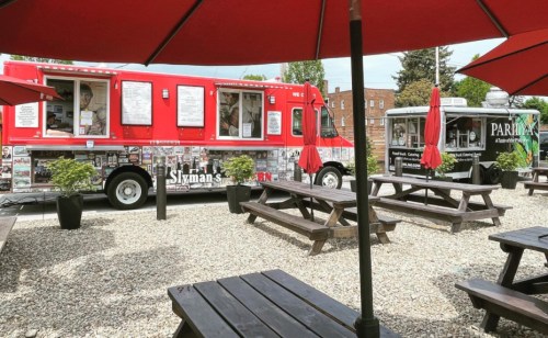 Two food trucks parked in a gravel area with picnic tables and red umbrellas. One truck is red, the other silver.