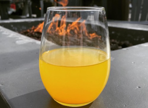 A clear glass filled with bright yellow liquid sits on a table near a fire pit with flames in the background.