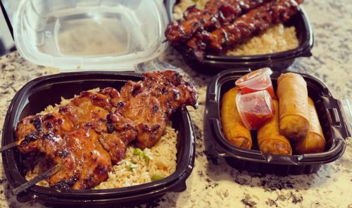 Two takeout containers with grilled skewers over rice and spring rolls with dipping sauce on a marble countertop.