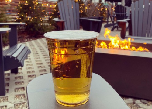 A clear plastic cup filled with golden beer sits on a table near a fire pit, with outdoor seating in the background.