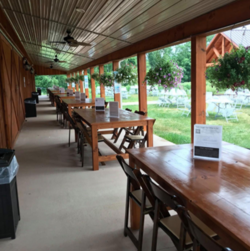 A covered outdoor dining area with long wooden tables, chairs, and hanging flower baskets, surrounded by greenery.