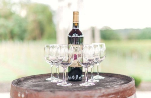 A bottle of red wine surrounded by eight wine glasses on a wooden barrel, set against a vineyard backdrop.