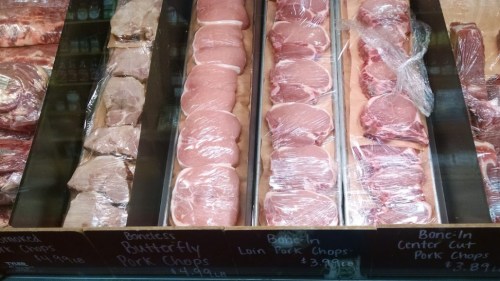 Display of various pork chops wrapped in plastic, labeled with prices, in a butcher shop.
