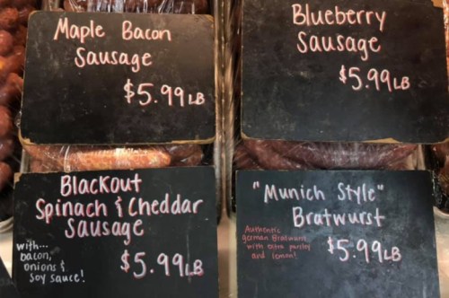 Four types of sausages displayed on chalkboard signs: Maple Bacon, Blueberry, Blackout Spinach & Cheddar, and Munich Style Bratwurst.
