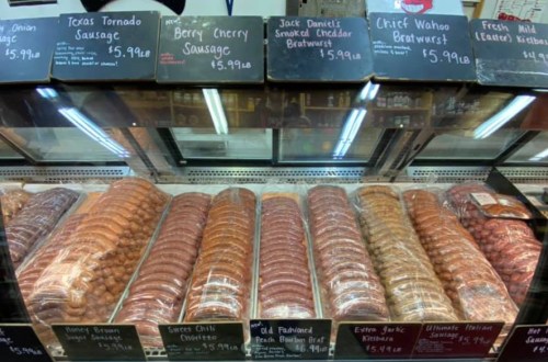 Display of various sausages and bratwursts in a butcher shop, labeled with prices and names.