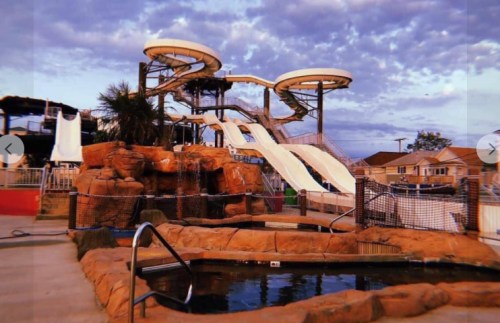 Water slides tower over a rocky landscape, with a small pool in the foreground and a cloudy sky above.