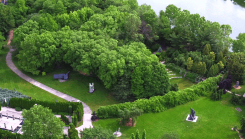Aerial view of a lush green park with walking paths, sculptures, and a serene water body in the background.