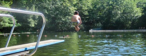 A child jumps off a diving board into a green, forested lake on a sunny day.