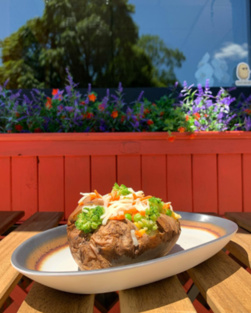 A baked potato topped with broccoli, shredded cheese, and mixed vegetables, set on a wooden table with flowers in the background.