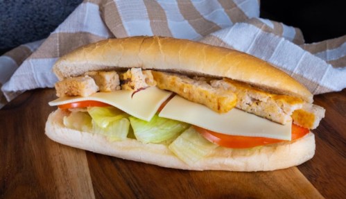 A sandwich on a wooden board, filled with lettuce, tomato, cheese, and sliced chicken, with a striped cloth in the background.