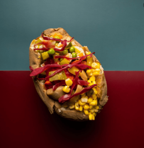 A baked potato topped with corn, green peas, and colorful pickled vegetables on a two-tone background.