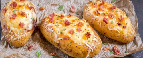 Three loaded baked potatoes topped with cheese, bacon bits, and herbs on a piece of parchment paper.