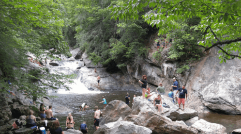 A scenic river with people swimming and relaxing on rocks, surrounded by lush greenery and a waterfall in the background.