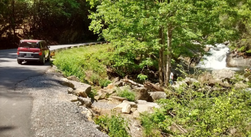 A red vehicle parked by a winding road next to a lush green area and a flowing creek with a small waterfall.
