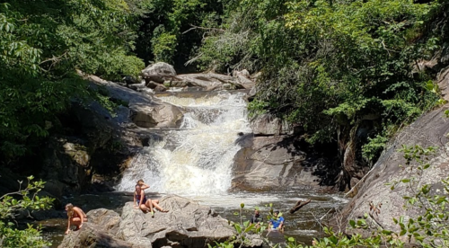 A scenic waterfall surrounded by lush greenery, with people relaxing on rocks near the water.