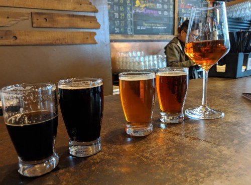 A row of four beer glasses, including dark and light beers, alongside a large wine glass on a wooden table.