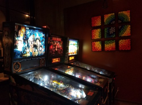 Three illuminated pinball machines in a dimly lit room, with colorful artwork on the wall behind them.