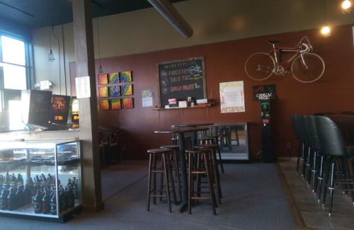 A cozy café interior with a bike on the wall, bar stools, a chalkboard menu, and a display of drinks.