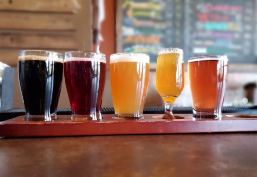 A flight of five different craft beers in glasses on a wooden paddle, with a blurred bar menu in the background.