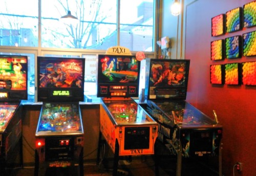 A row of vintage pinball machines in a brightly lit arcade, with colorful artwork and a vibrant wall display.