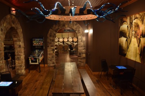 Interior of a rustic venue featuring wooden tables, stone arches, and a unique chandelier, with barrels in the background.