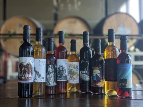 A collection of wine bottles with artistic labels displayed on a wooden table, with barrels in the background.