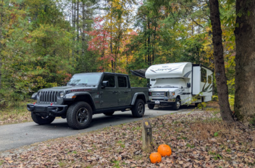 A Jeep Gladiator parked beside a camper on a forested road, with autumn foliage and two pumpkins nearby.