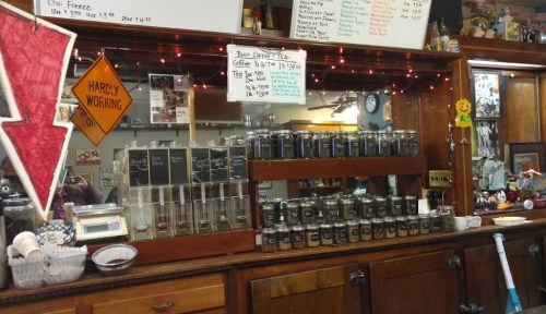 A wooden counter displaying jars of coffee and tea, with a menu board and decorative lights in a cozy café setting.