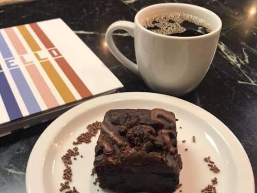 A cup of coffee beside a plate with a chocolate brownie topped with chocolate drizzle and sprinkles, on a marble table.