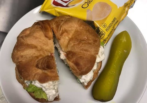 A plate with a halved croissant sandwich filled with chicken salad, a pickle, and a bag of Lay's potato chips.