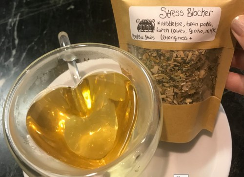 A heart-shaped glass cup filled with golden herbal tea next to a packet labeled "Stress Blocker" with herbal ingredients.