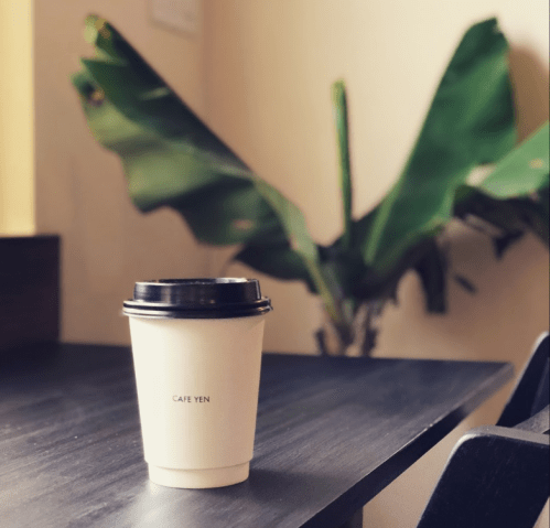 A coffee cup labeled "CAFE YEN" sits on a table, with a large green plant in the background.