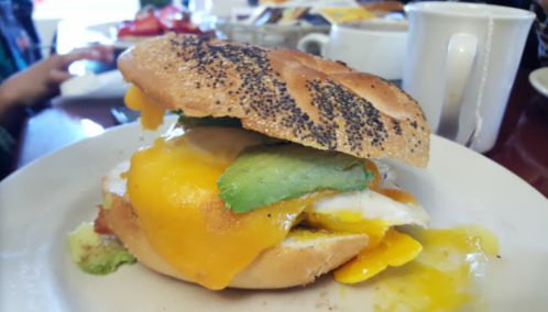 A poppy seed bagel sandwich with melted cheese, avocado, and a fried egg, served on a white plate.