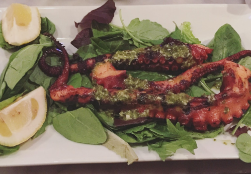 Grilled octopus served on a bed of greens, garnished with sauce and lemon wedges.