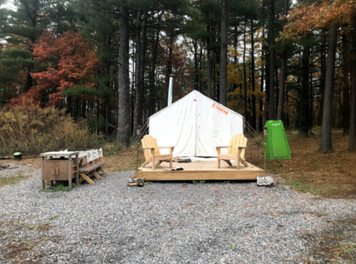 A cozy camping setup with a white tent, wooden deck, two chairs, and a green tent, surrounded by trees in autumn.