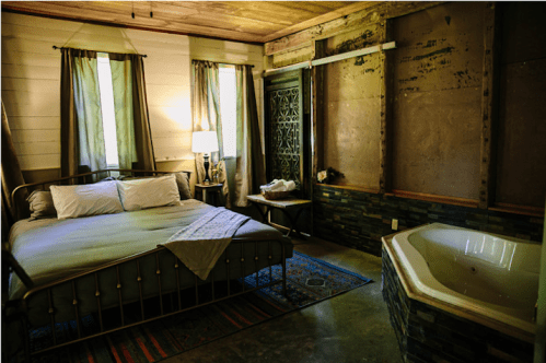 Cozy bedroom with a metal bed, soft lighting, and a jacuzzi tub, featuring rustic decor and green curtains.