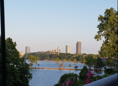 City skyline reflected in a calm lake, surrounded by greenery and trees under a clear blue sky.