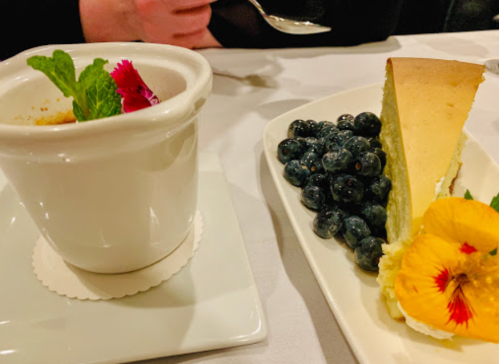 A dessert plate featuring cheesecake, blueberries, and a mint garnish, alongside a small cup with a flower.