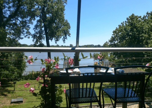 A scenic view from a balcony overlooking a river, with flowers and chairs in the foreground under a clear blue sky.