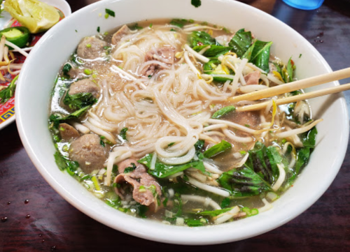 A bowl of noodle soup with meat, herbs, and bean sprouts, served with chopsticks on the side.
