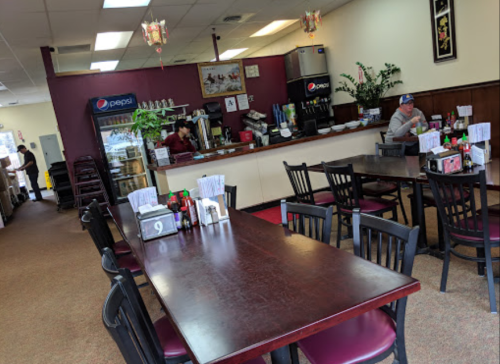 A cozy restaurant interior with tables, a counter, and a few patrons. Decor includes plants and menu items on tables.