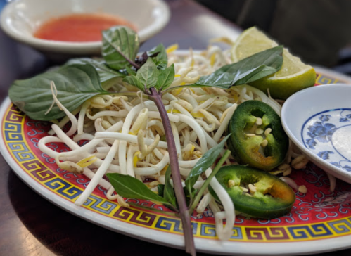 A colorful plate of fresh bean sprouts, basil, lime wedges, and sliced jalapeños, with a small bowl of sauce in the background.