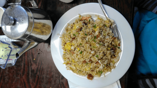 A plate of fried rice with shrimp, vegetables, and a fork, next to a teapot on a wooden table.