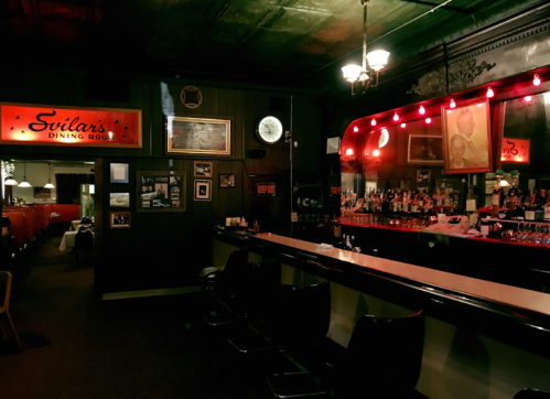 A dimly lit bar area with a long counter, vintage decor, and red lights, creating a cozy atmosphere.