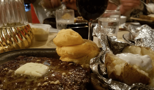 A close-up of a sizzling steak with butter, a baked potato in foil, and a soft roll, with drinks in the background.