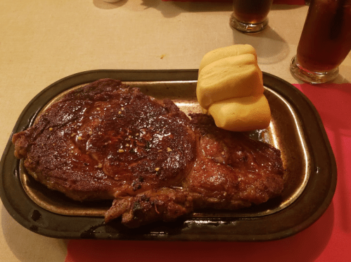 A juicy steak on a plate with two soft dinner rolls, accompanied by glasses of iced tea in the background.