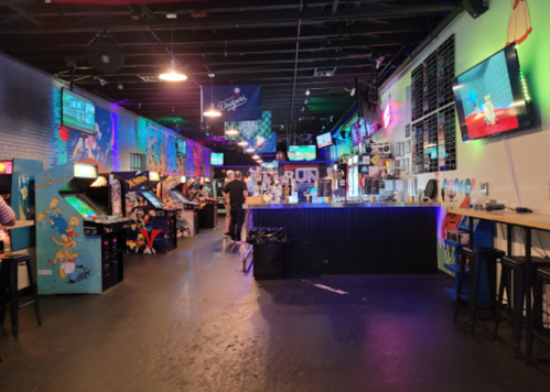 A vibrant arcade bar with colorful lights, vintage arcade machines, and a bar area, filled with patrons enjoying the space.